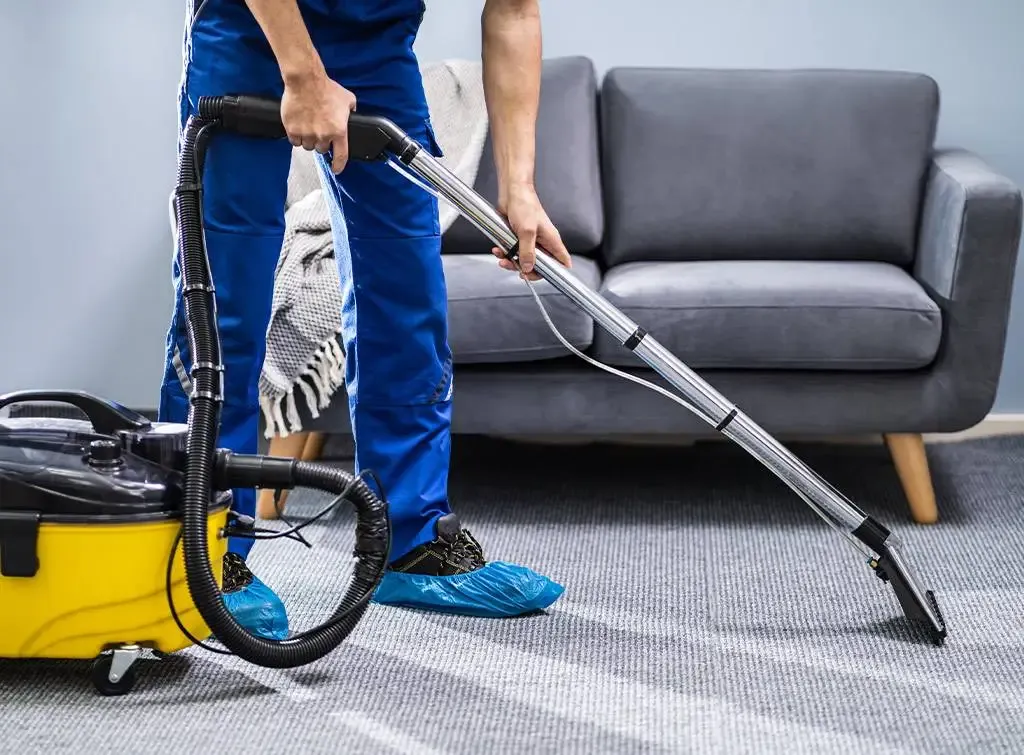 A person in shoe covers vacuuming a grey carpet beside a sofa