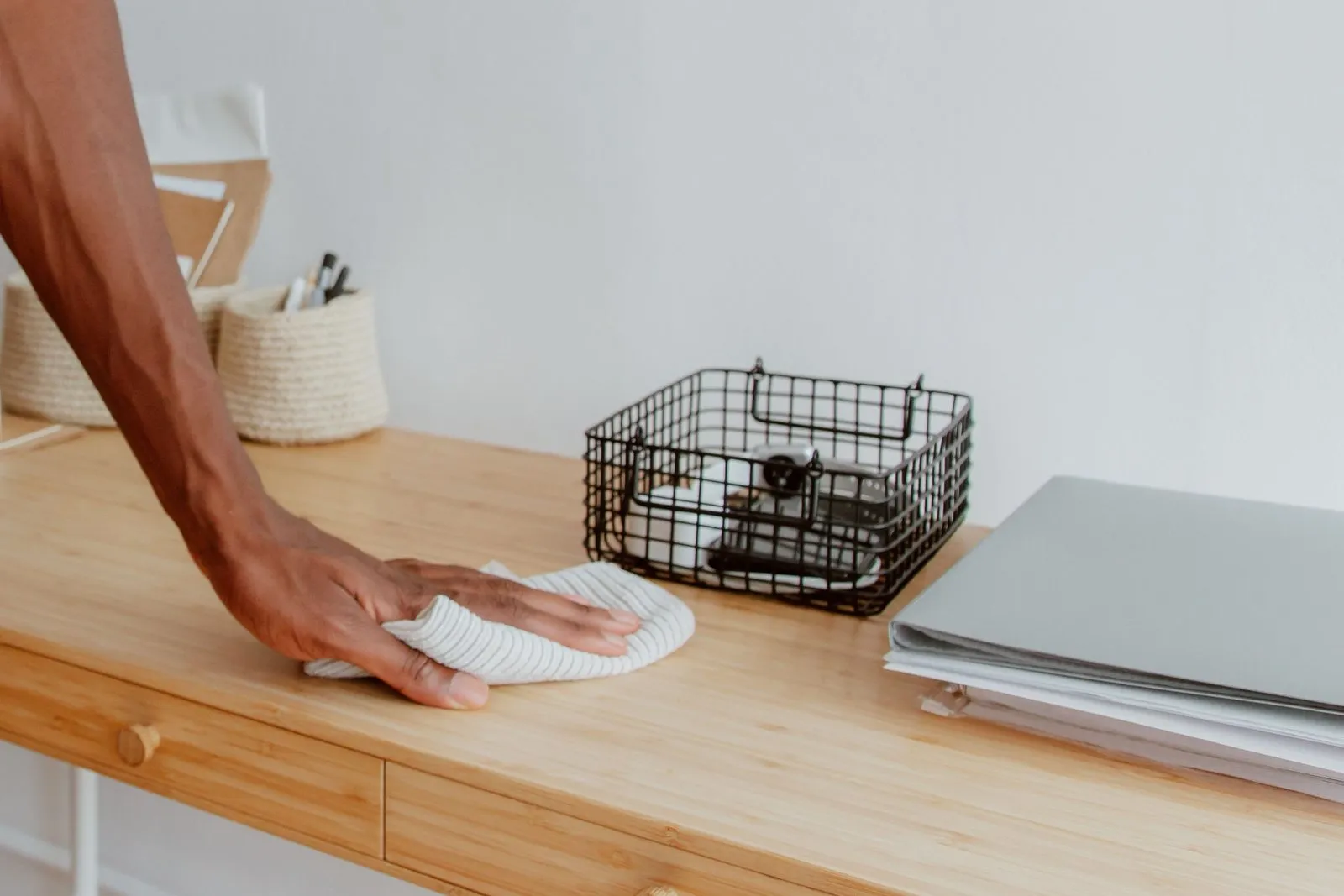A hand wiping a wooden desk with a cloth, beside a wire basket and a closed laptop