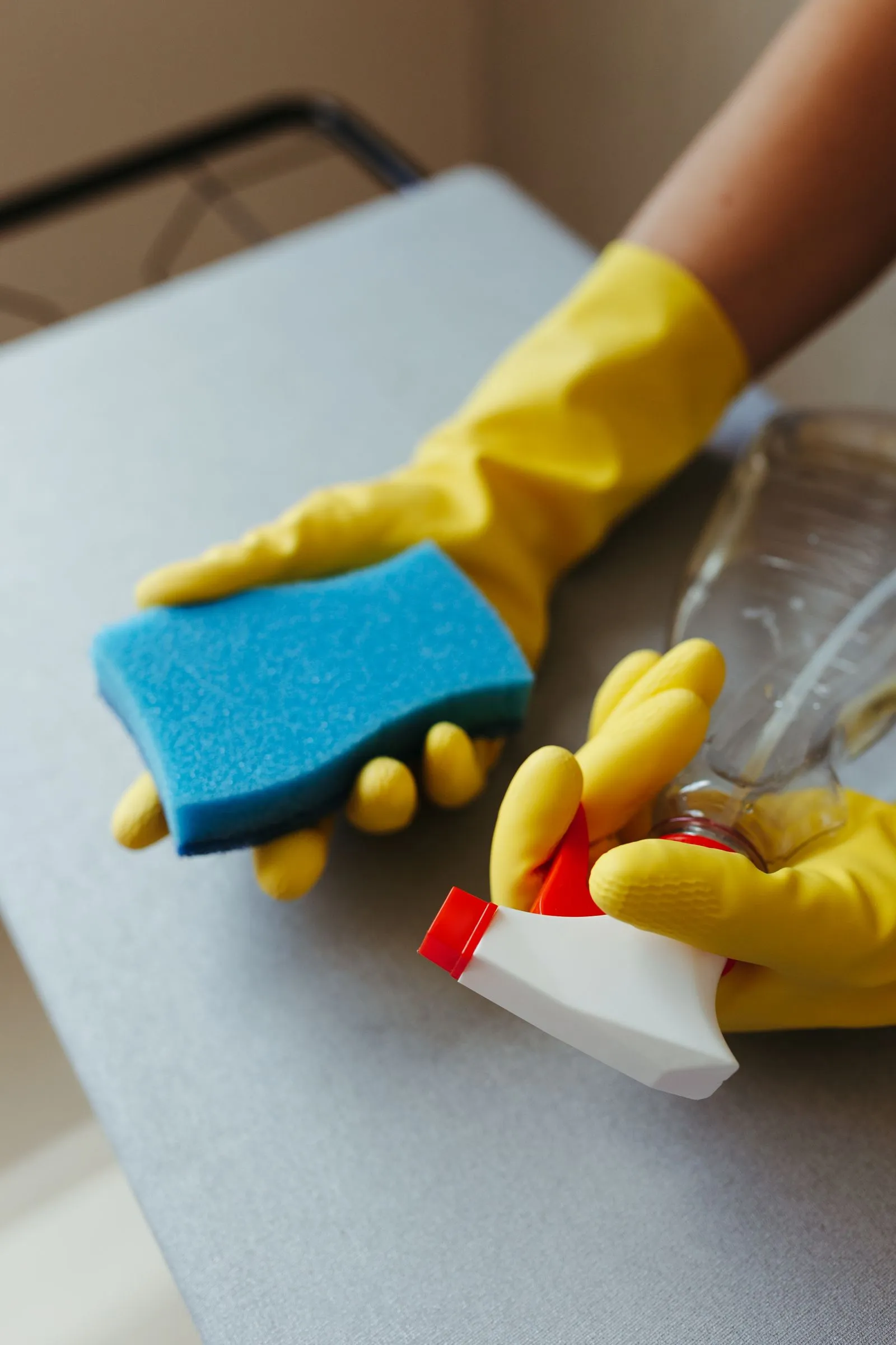 Hands in yellow rubber gloves holding a blue sponge and a spray bottle over a grey surface