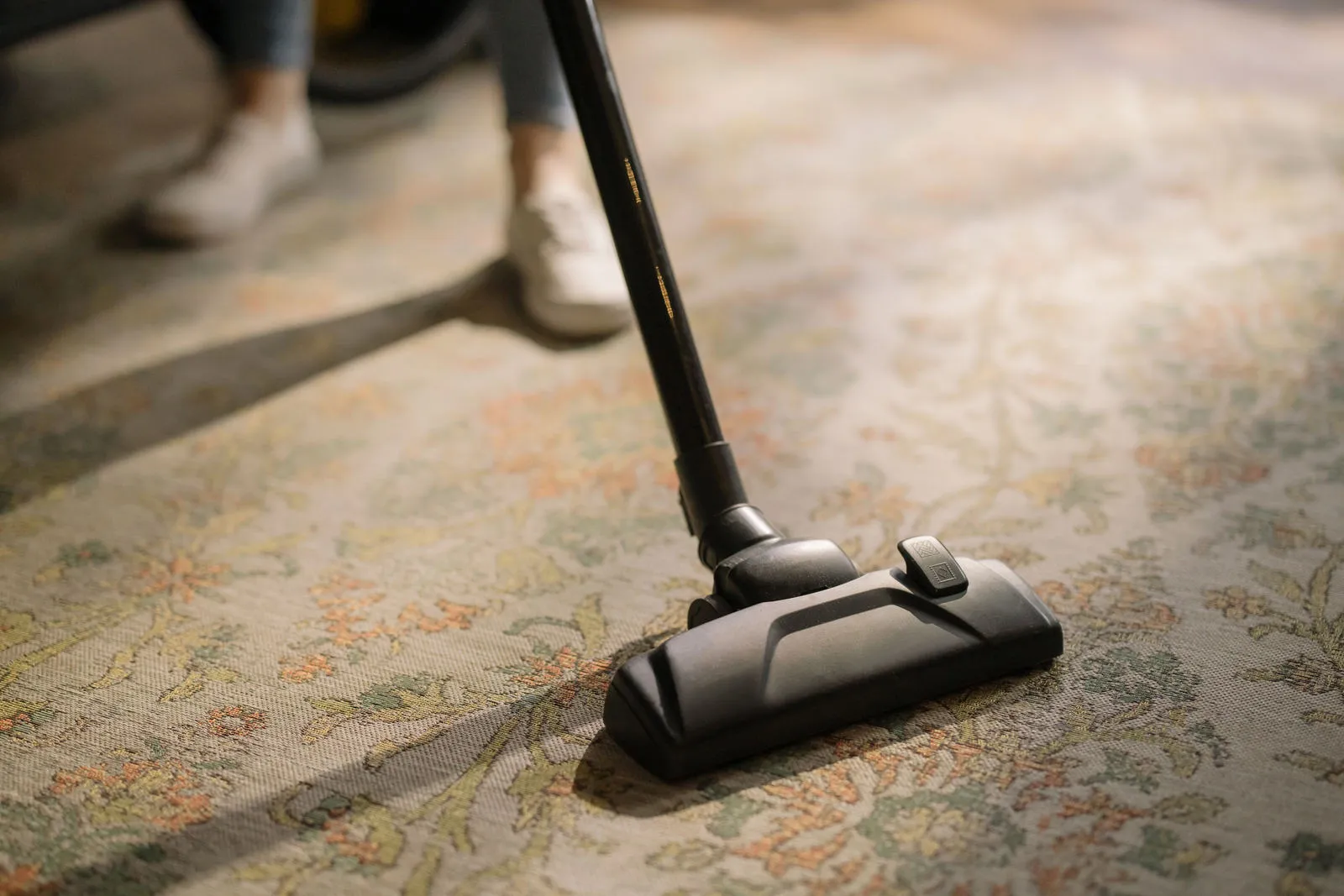 A vacuum cleaner head on a patterned rug, with a person in white trainers behind it