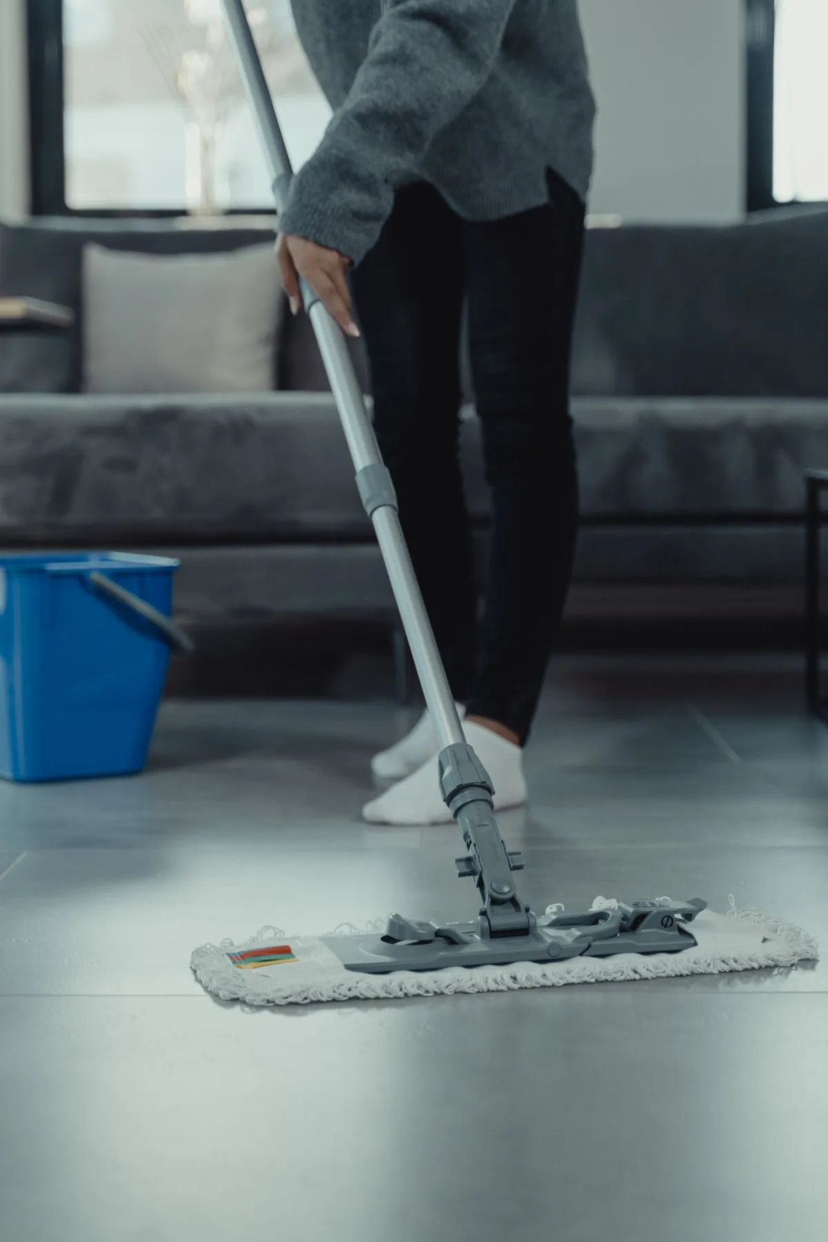 A person in white socks mopping a grey tiled living room floor beside a sofa and a blue bucket