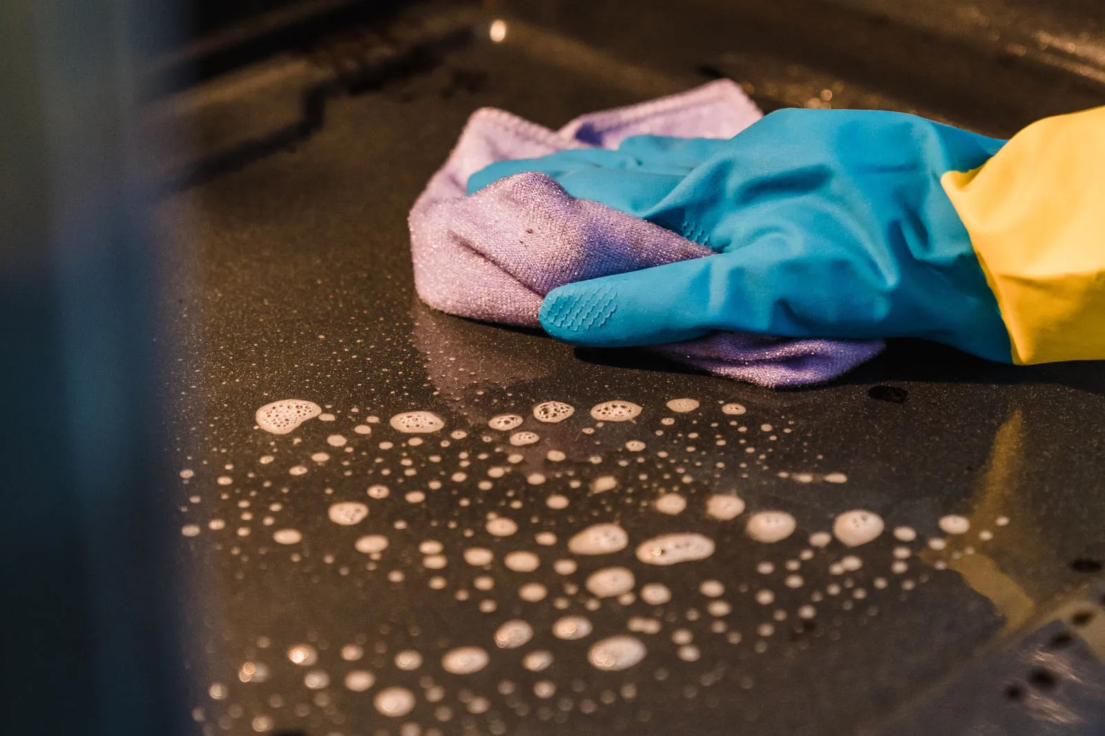 A hand in a rubber glove wiping the inside of an oven with a cloth and soapy water